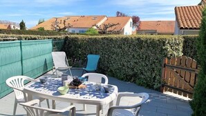 An outdoor patio with a round table and white plastic chairs, surrounded by a green fence and a wooden gate. - La Palmyre - Le HAMEAU de TALARIS - TERRACED HOUSE - SHARED POOL (LA PALMYRE)