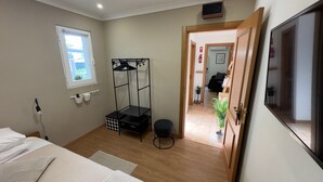 Une chambre à coucher comprenant un lit, une armoire en métal noir, un petit tabouret, une fenêtre et une porte donnant sur une autre pièce.