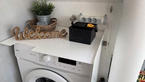 Un lave-linge à chargement frontal doté d’un panneau de commande, d’une pancarte de bienvenue, d’une plante en pot et d’un panier contenant divers objets.