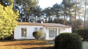 Une petite maison blanche avec un toit en tuiles, entourée de pins et d’une pelouse bien entretenue.