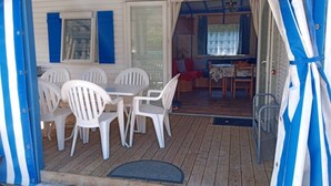 Una puerta abierta a un espacio exterior cubierto con terraza de madera, sillas de plástico blanco y una mesa.