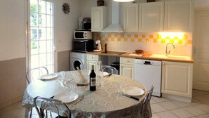 Cozinha compacta com armários brancos, micro-ondas, máquina de café e pia. Há uma mesa de jantar posta com pratos, uma garrafa de vinho e talheres.