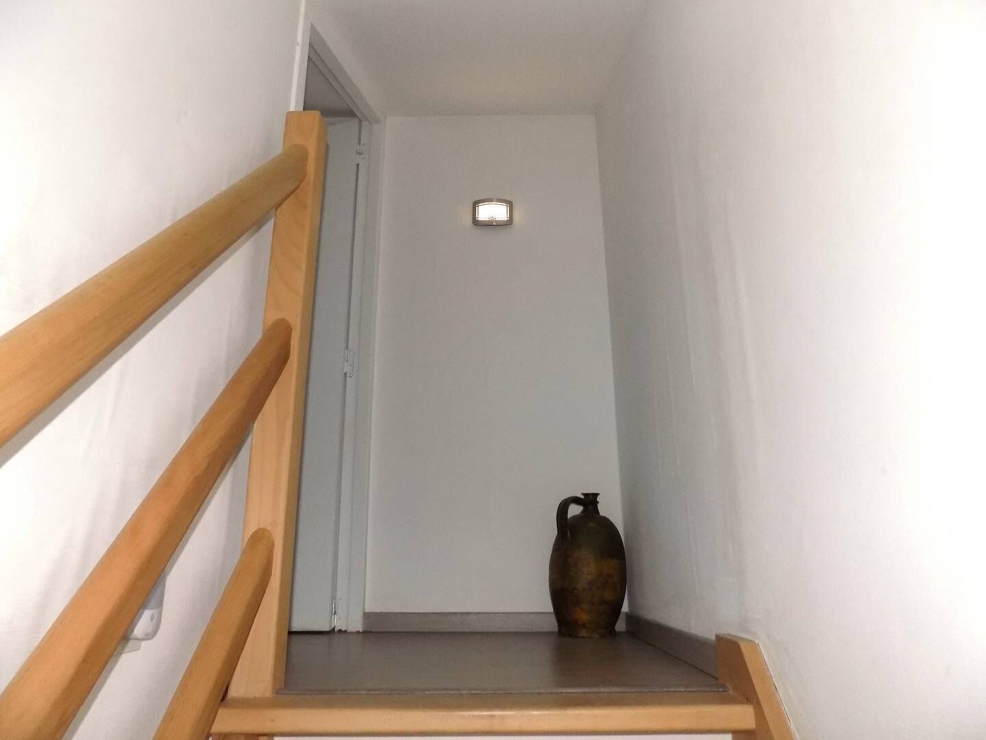 A narrow stairway with wooden handrails leading to a door. A small, dark-colored vase is placed on the landing.