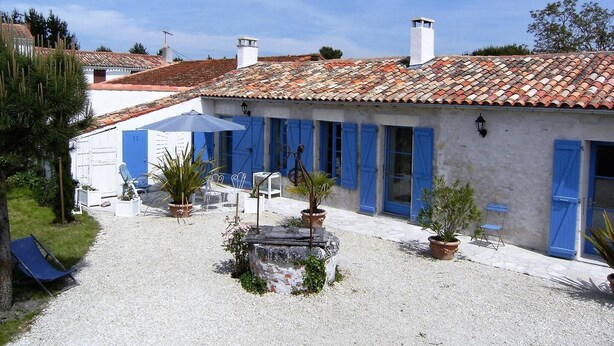 A courtyard with a white building featuring blue shutters and a tiled roof. There are potted plants and outdoor furniture. - Les Mathes La Palmyre - CHARMING CHARENTAISE HOUSE - PEACEFUL - COUNTRYSIDE (LES MATHES)