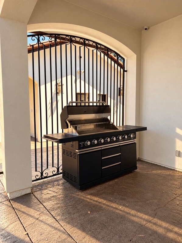 Una parrilla exterior negra con cuatro quemadores, un estante lateral y una chimenea, ubicada en un patio cubierto con una reja metálica al fondo.
