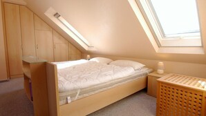 Ein Schlafzimmer mit schrägverlaufender Decke, einem Bett mit weißer Bettwäsche, einem Nachttisch aus Holz mit Lampe und einem Wäschekorb.