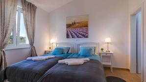 Een tweepersoonsbed met een donkerblauwe en grijze dekbedovertrek, twee nachtkastjes met lampen, een schilderij van een veld en een raam met gordijnen.