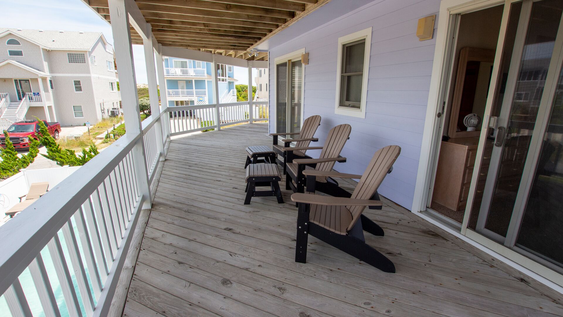 A covered outdoor deck with wooden chairs and a table, a sliding glass door, and a view of neighboring buildings.