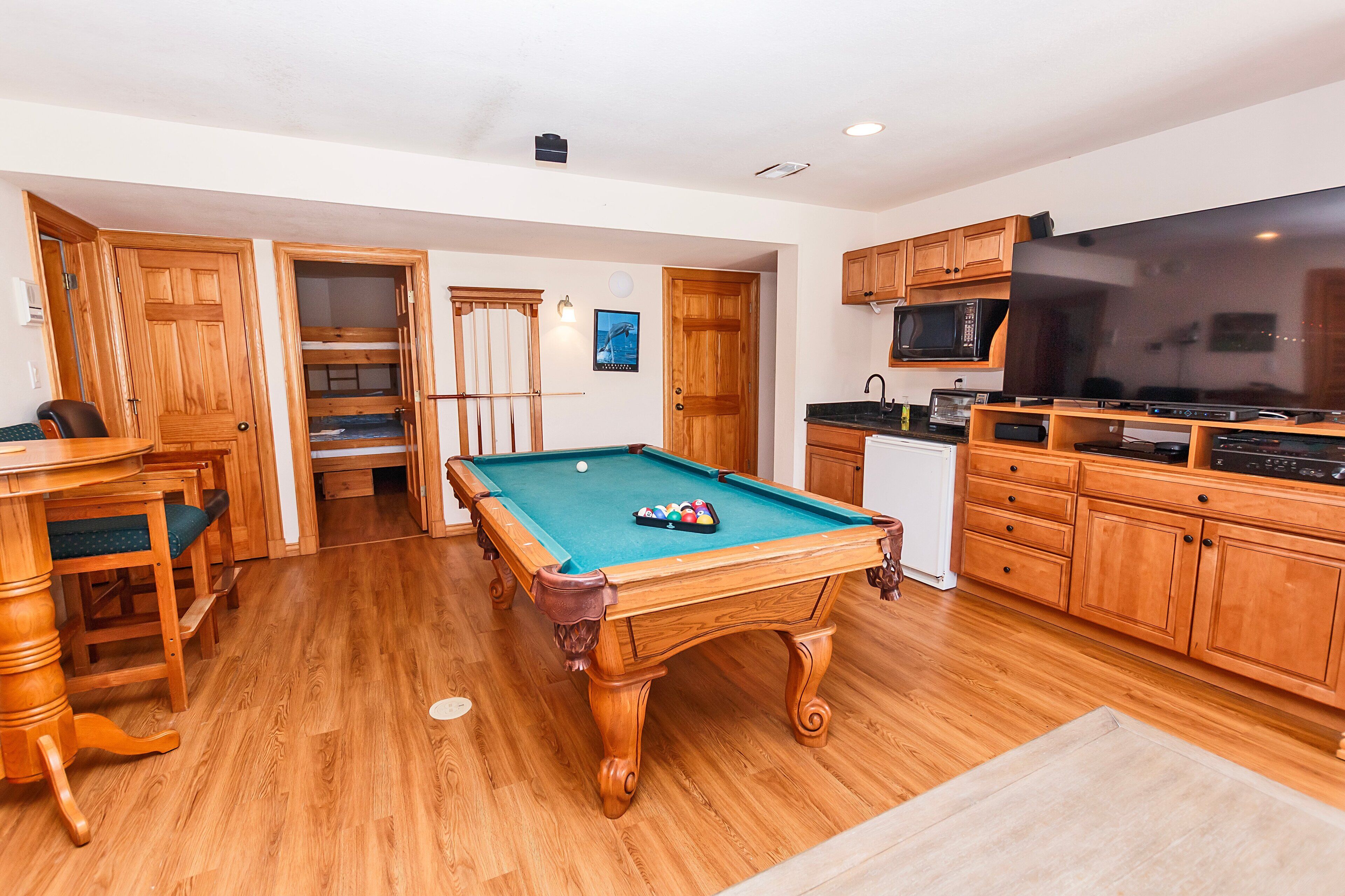 A pool table in a room with wooden flooring, wooden cabinets, a microwave, and a television.