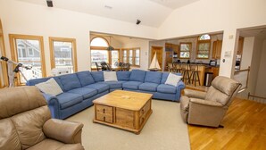 A spacious living room with a large blue sectional sofa, a wooden coffee table, and leather armchairs. There is a kitchen area with wooden cabinets and a dining area with chairs. - OCEANFRONT, Private Pool, Ocean Views, Shared Beach Walkway, BU10, Summer Paradise (Corolla)