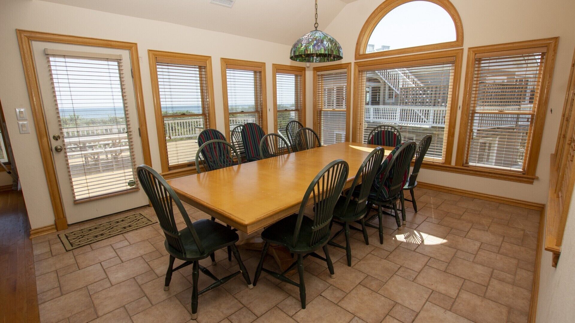 A dining room with a large wooden table and chairs, large windows with blinds, and a hanging light fixture.