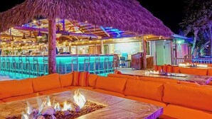 Un área junto a la piscina con techo de paja, asientos anaranjados y una chimenea.