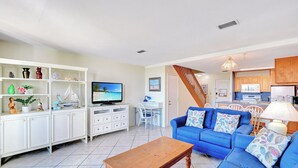 Una sala de estar con un sofá azul, una mesa de centro de madera, una estantería blanca y un televisor.