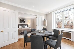 A hotel room with a bed, a dining table with chairs, a large window with a view of buildings, and a door leading to another room. - Stylish apartment with terrace in Mayfair (London)