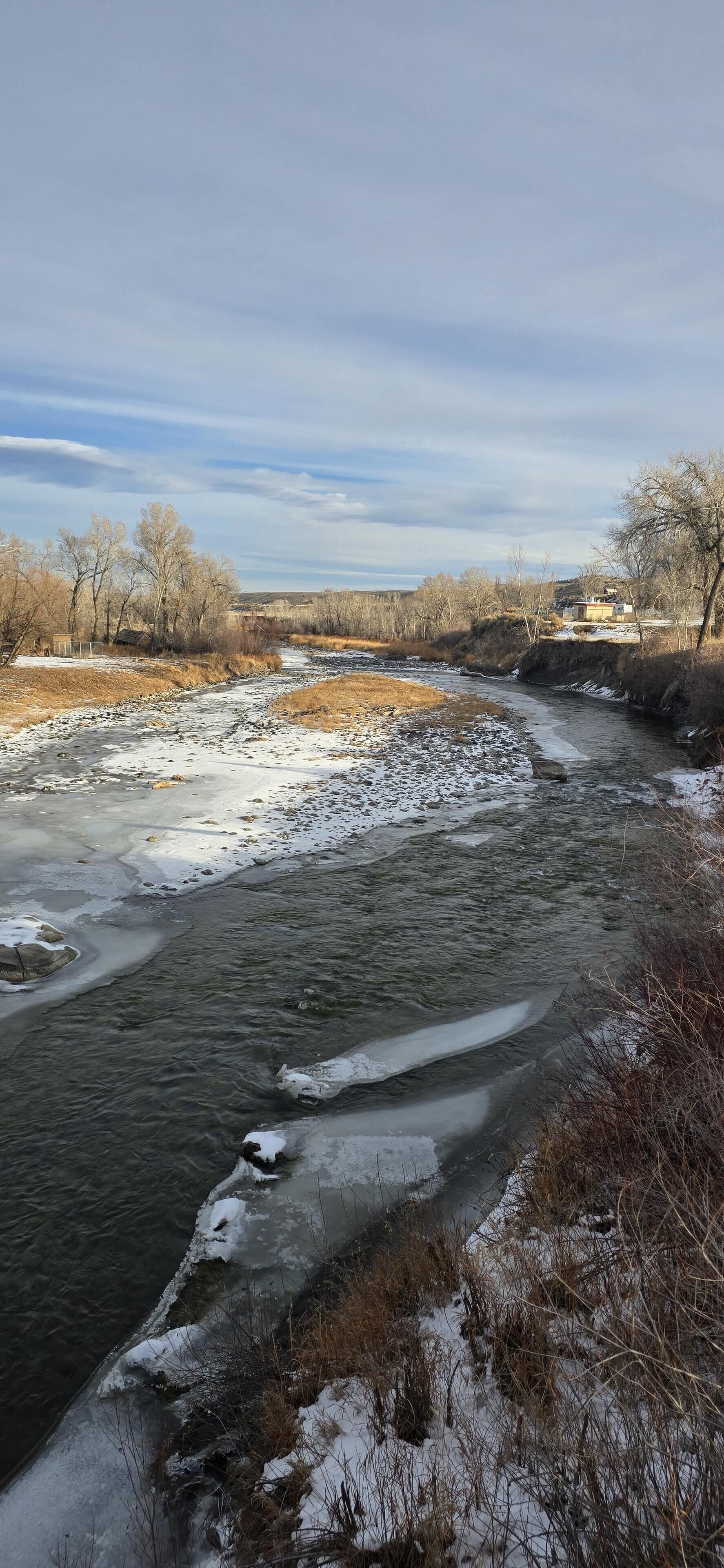 Une rivière dont les berges sont partiellement recouvertes de glace, entourée d’arbres sans feuilles et sous un ciel dégagé.