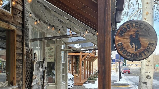 A wooden deck with string lights, a sign reading "Meteetse Chocolate," and a view of a snowy street. - Stargazer Bunkhouse- cozy cabin, basecamp for adventures in Yellowstone and beyond (Meeteetse)
