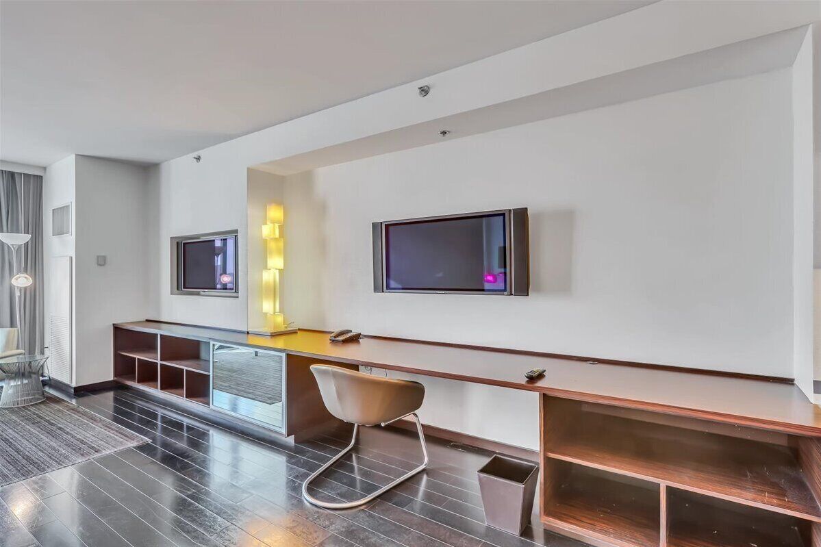 A modern hotel room with a large flat-screen TV mounted on the wall, a desk with a chair, and a built-in bench along the wall.