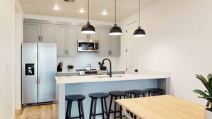 A modern kitchen with white cabinets, a stainless steel refrigerator, a microwave, and a sink. There are three black bar stools and a wooden dining table. - Desert Memories #76 by RedAwning (St. George)