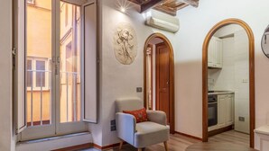 Une pièce dotée d’une porte-fenêtre coulissante, d’un petit fauteuil avec un coussin, d’une table en bois et d’une cuisine visible à travers une arche.