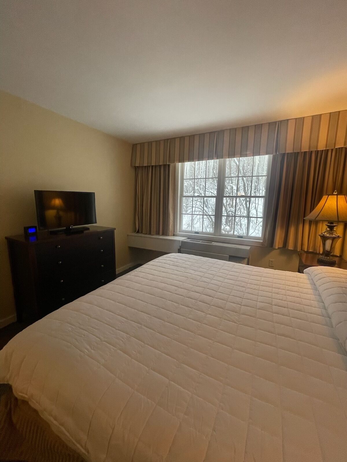 Ein Hotelzimmer mit einem Bett, einem Fernseher auf einem Schminktisch, einem Fenster mit Vorhängen und einer Lampe.