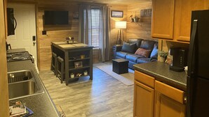 A compact kitchen with a stove, sink, and countertop. There is a wooden dining table with chairs, a sofa, and a small coffee table. The room has wooden walls and a ceiling with recessed lighting. - Grand Rivers, KY - Unit A - Cozy getaway with Cabin Charm near KY & Barkley Lake (Grand Rivers)
