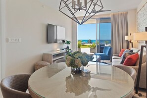 Een moderne hotelkamer met een glazen eettafel, een flatscreen televisie, een bank met kleurrijke kussens en een balkon met uitzicht op de oceaan.
