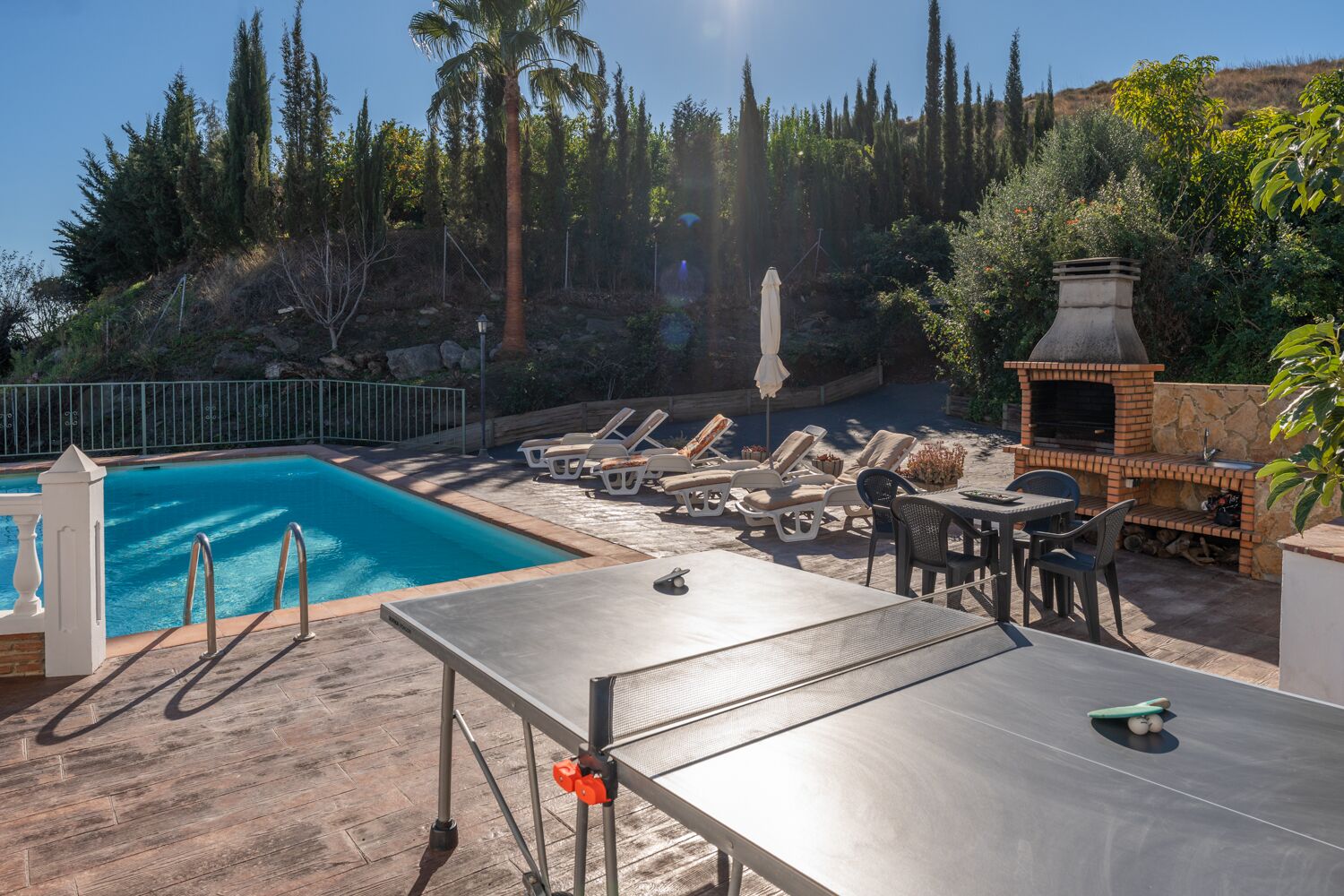 Zona de piscina amb gandules, una barbacoa i una taula.