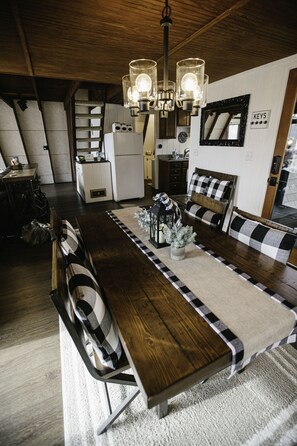 A dining area with a long wooden table, black and white checkered chairs, a chandelier, and a kitchen in the background.