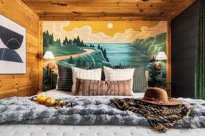 A cozy bedroom with a mural of a landscape, a bed with patterned pillows, a fluffy blanket, and a hat on the bed.