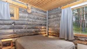 Een slaapkamer in een blokhut met een bed, twee bedlampen, een raam met gordijnen en uitzicht op een bos.