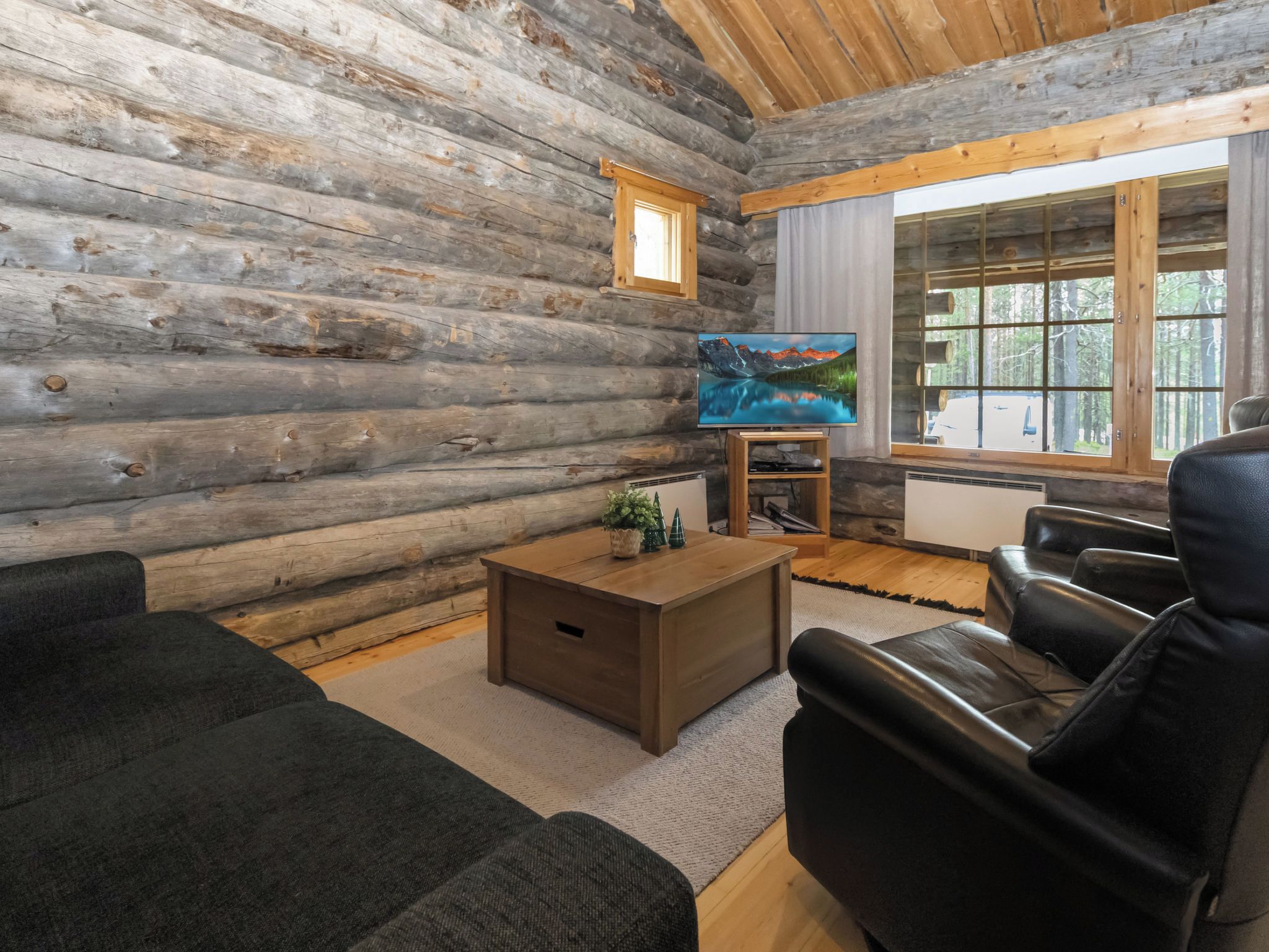 A log cabin interior with a wooden floor, a sofa, a coffee table, a television, and a window with a view of trees.