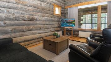 A log cabin interior with a wooden floor, a sofa, a coffee table, a television, and a window with a view of trees.