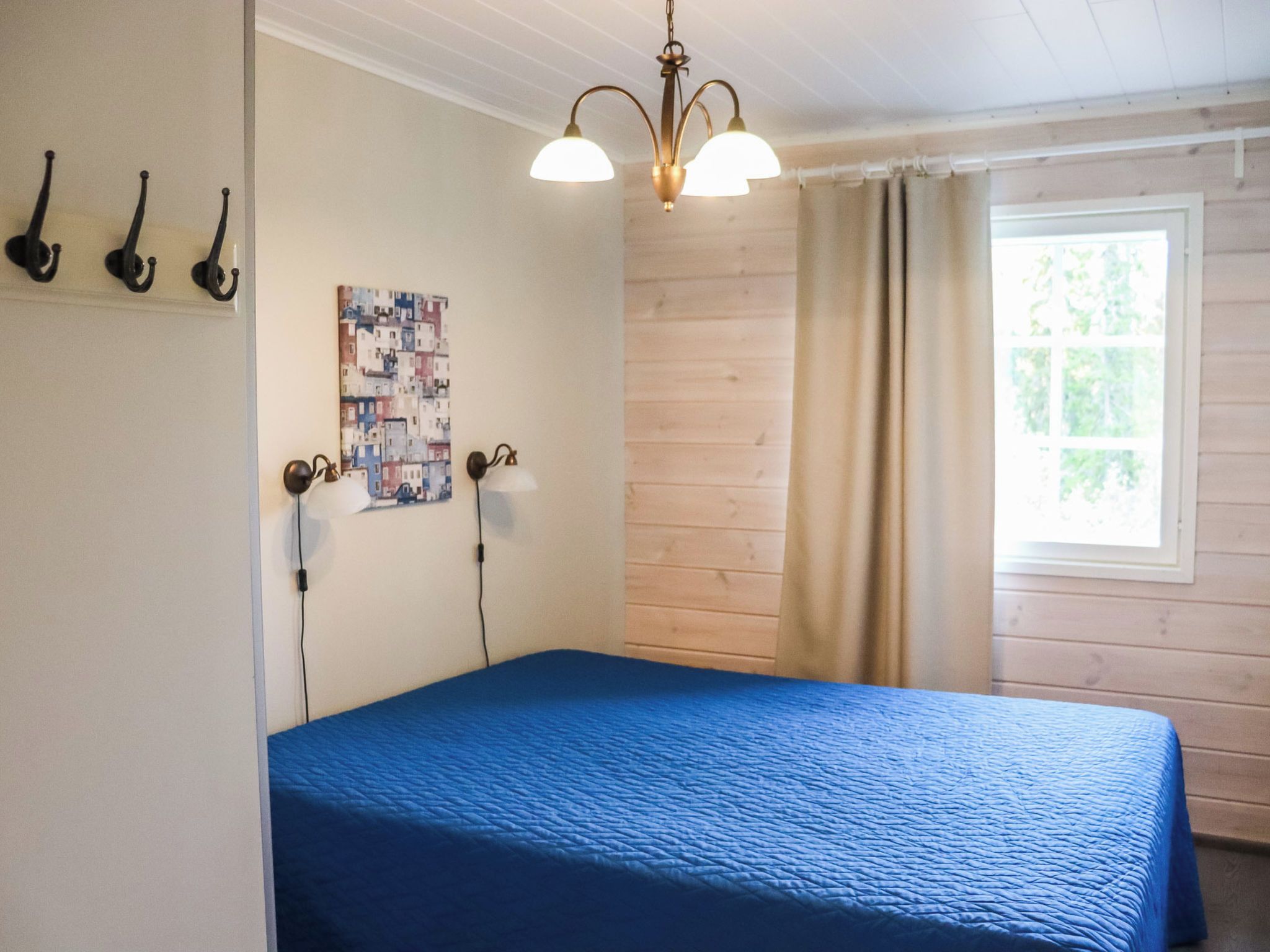 Een kamer met een bed, een wandlamp, een schilderij en een raam met gordijnen.