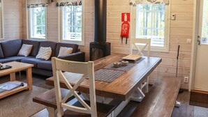 Interior acogedor de una cabaña con sofá, mesa de madera y sillas. Se ve un horno negro y ventanas con cortinas.