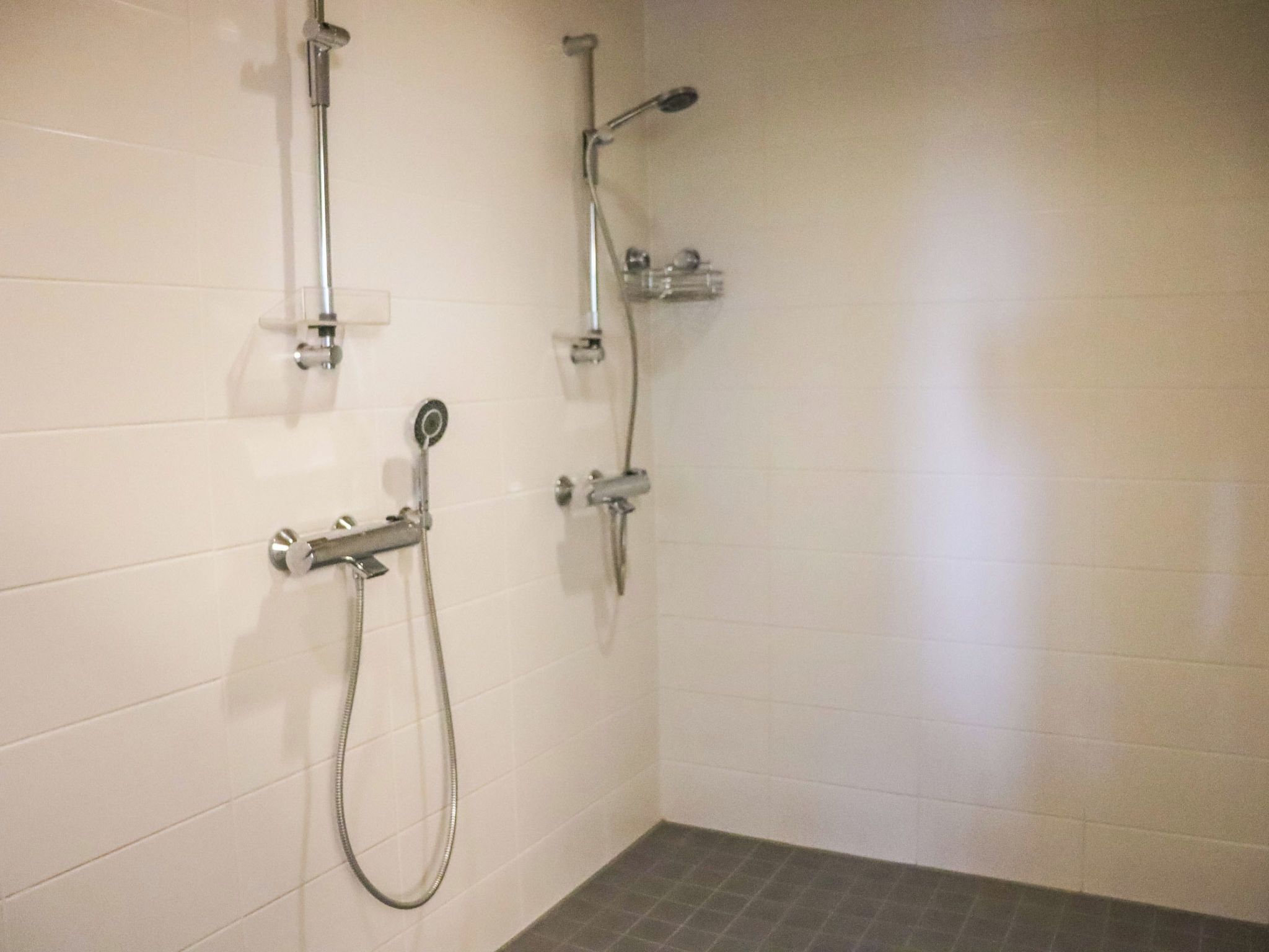 A bathroom with white tiled walls, a shower with a handheld showerhead, and a floor with dark tiles.