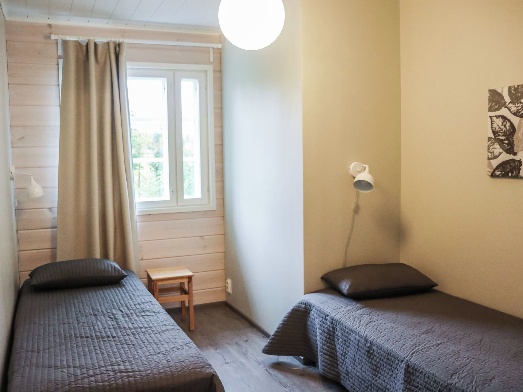 A room with two beds, a window with curtains, a small table, a wall-mounted lamp, and a wall art piece.