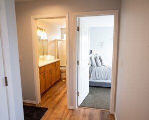 Un couloir étroit mène à une salle de bain équipée de toilettes et d’une douche, ainsi qu’à une chambre comprenant un lit et une table de chevet.