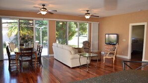 A spacious living room with hardwood flooring, a white sofa, a dining table with chairs, a TV, and a ceiling fan. - Charming Lakeview House, 15 minute drive to downtown Naples beaches and 5th Ave. (Naples)