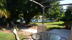 An outdoor patio with a round glass table and chairs, surrounded by a covered area with a view of a lake and trees. - Charming Lakeview House, 15 minute drive to downtown Naples beaches and 5th Ave. (Naples)