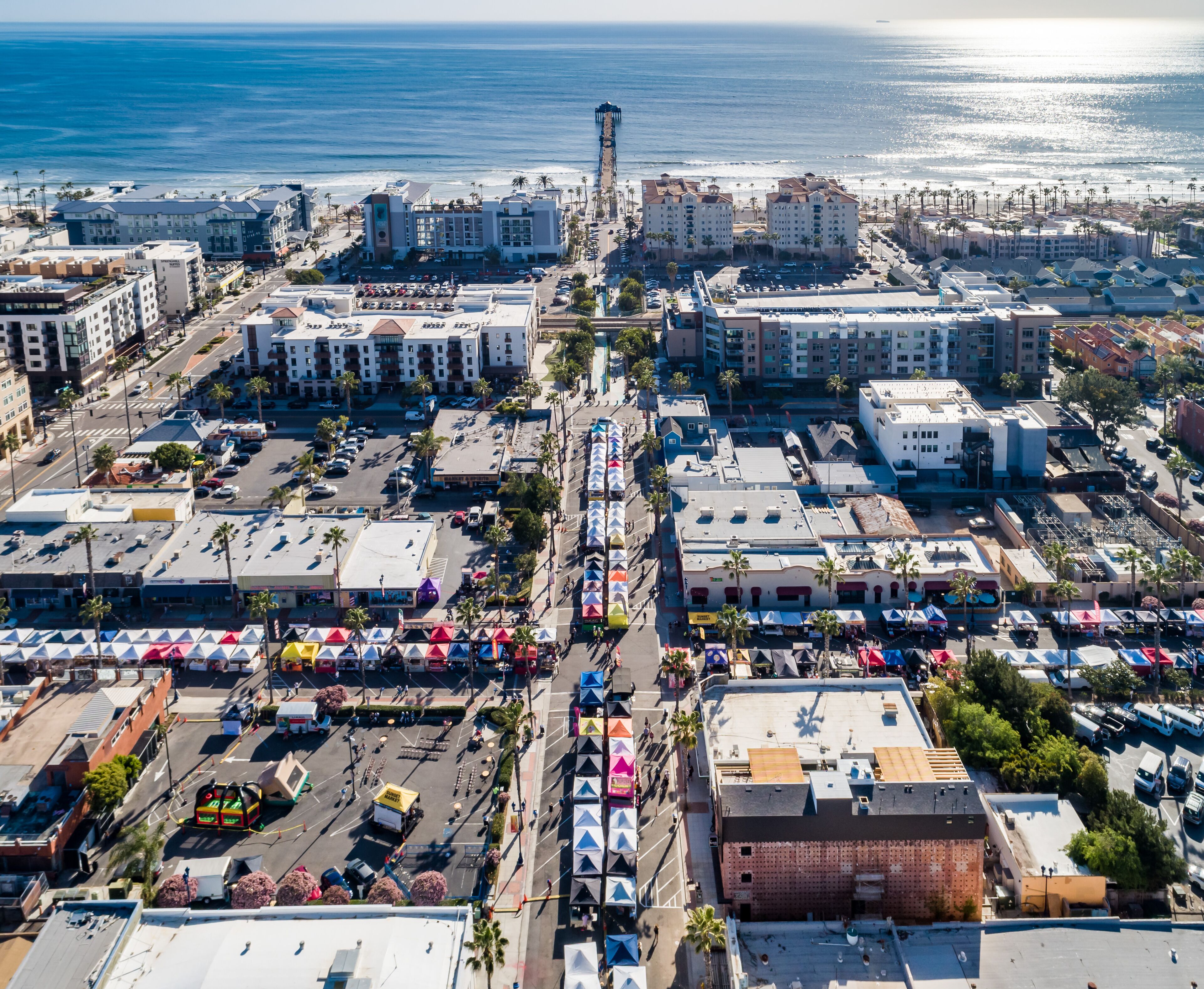An aerial view of a coastal city with buildings, a pier extending into the ocean, and a parking lot with colorful tents.