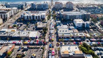 An aerial view of a coastal city with buildings, a pier extending into the ocean, and a parking lot with colorful tents.