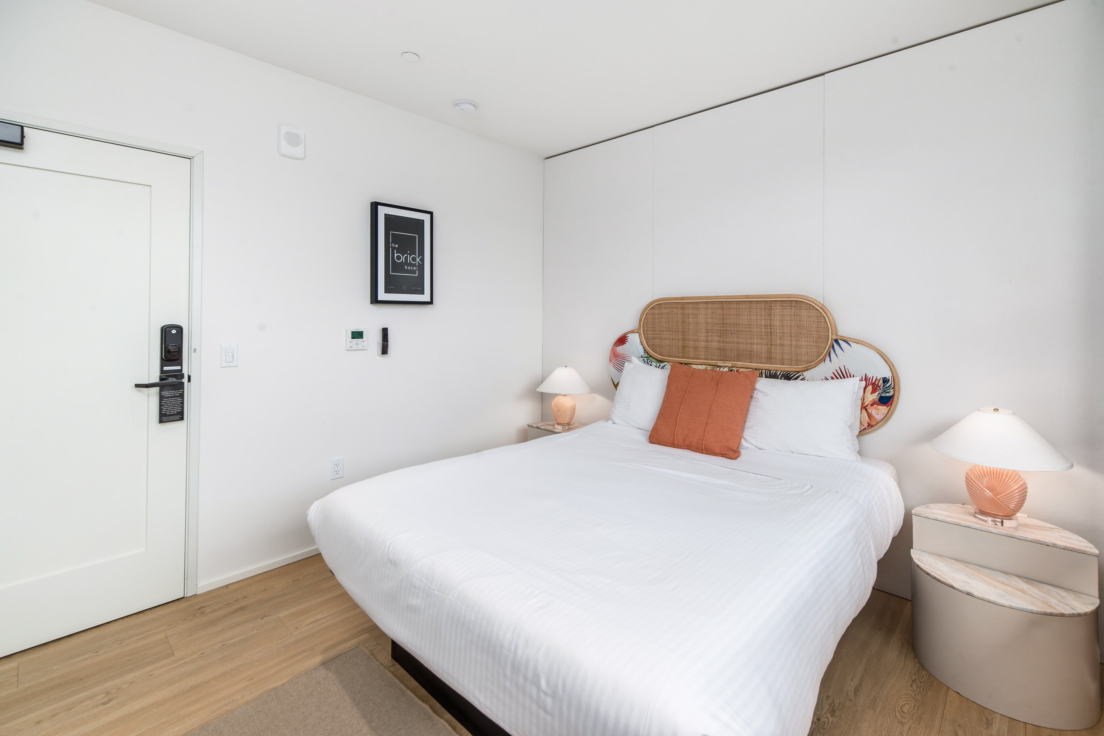 Een slaapkamer met een wit bed, twee nachtkastjes met lampen, een deur, een lijst met een afbeelding aan de muur en een houten vloer.