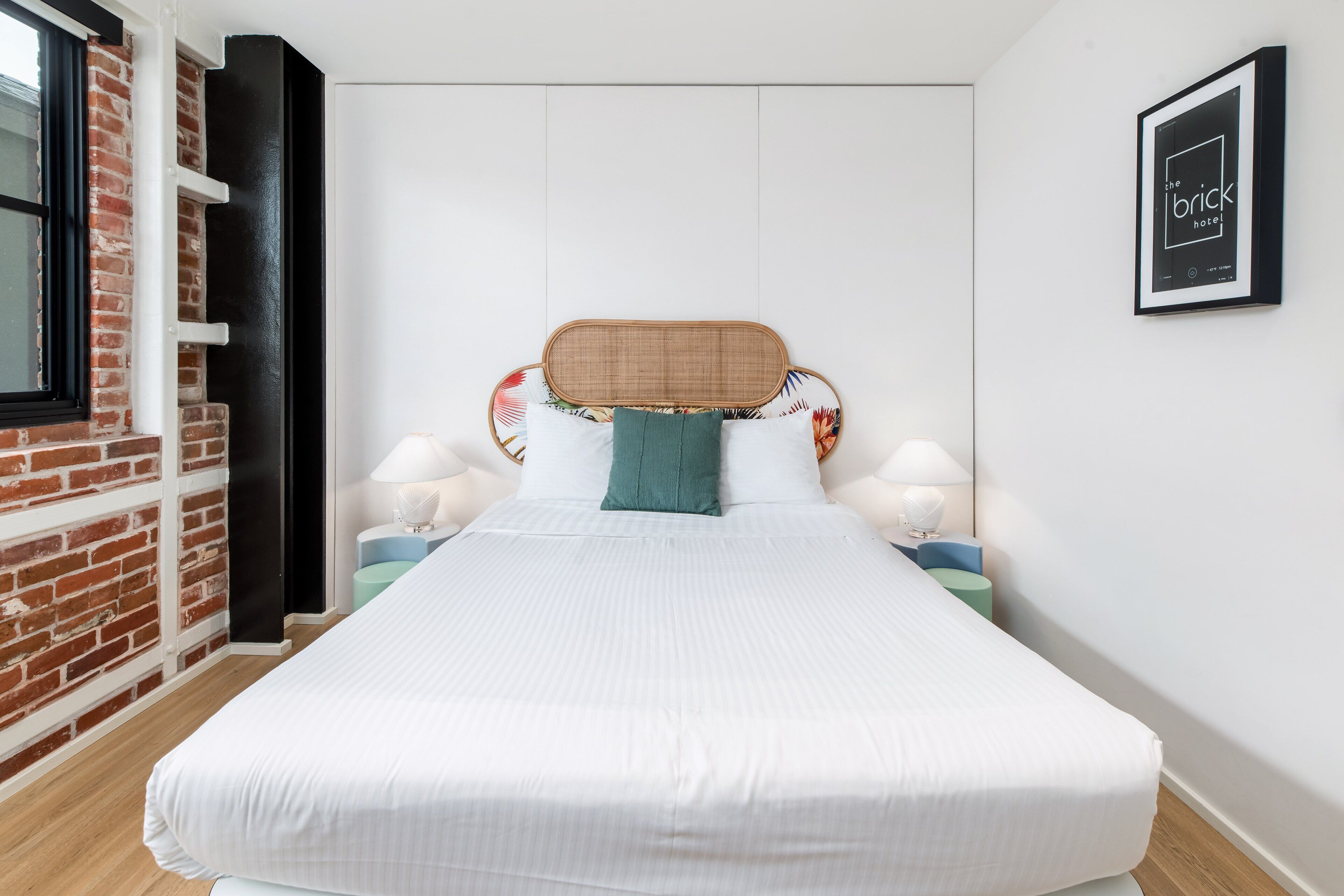 A modern hotel room with a brick accent wall, a large bed with white bedding, a bamboo headboard, and two bedside tables with lamps.