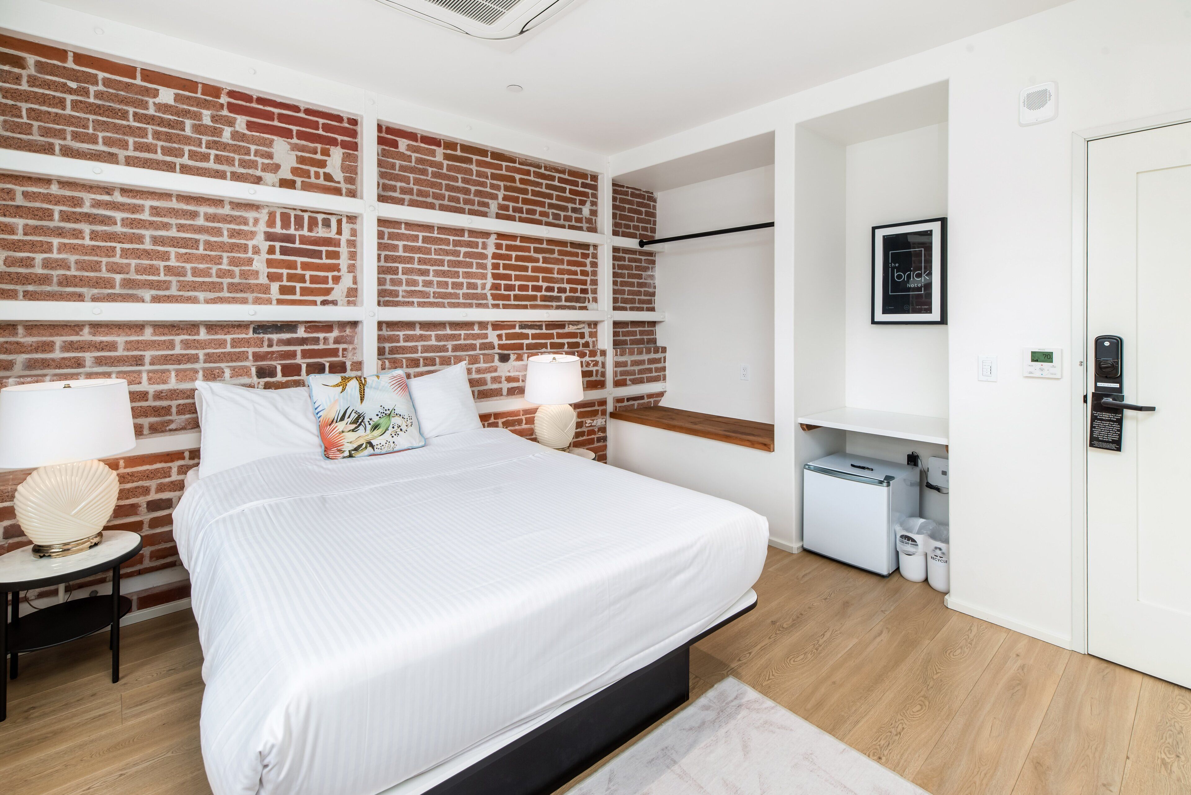 A modern bedroom with a brick-patterned wall, a large bed with white bedding, two bedside tables with lamps, a small white cabinet, and a door with a lock.