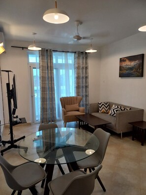 A modern living room with a glass-top dining table, a sofa, and a chair. There is a ceiling fan, a TV stand, and a painting on the wall.