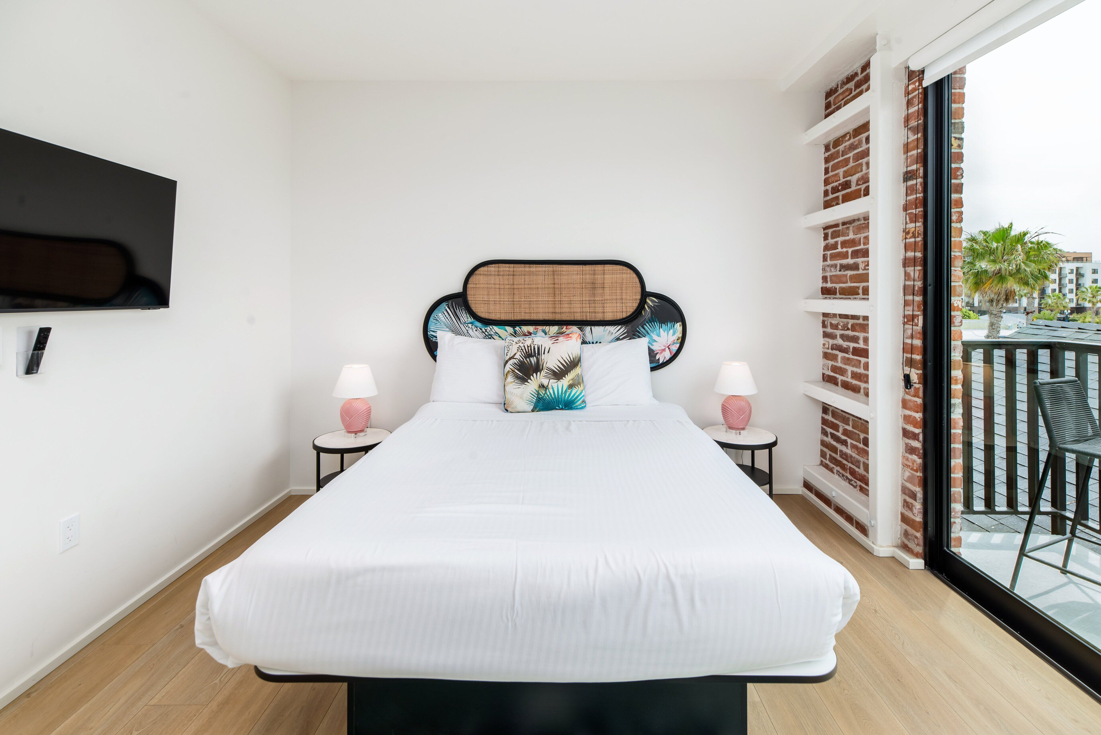 Un dormitorio con una cama grande, un televisor montado en la pared, dos mesitas de noche con lámparas y un balcón con una silla.