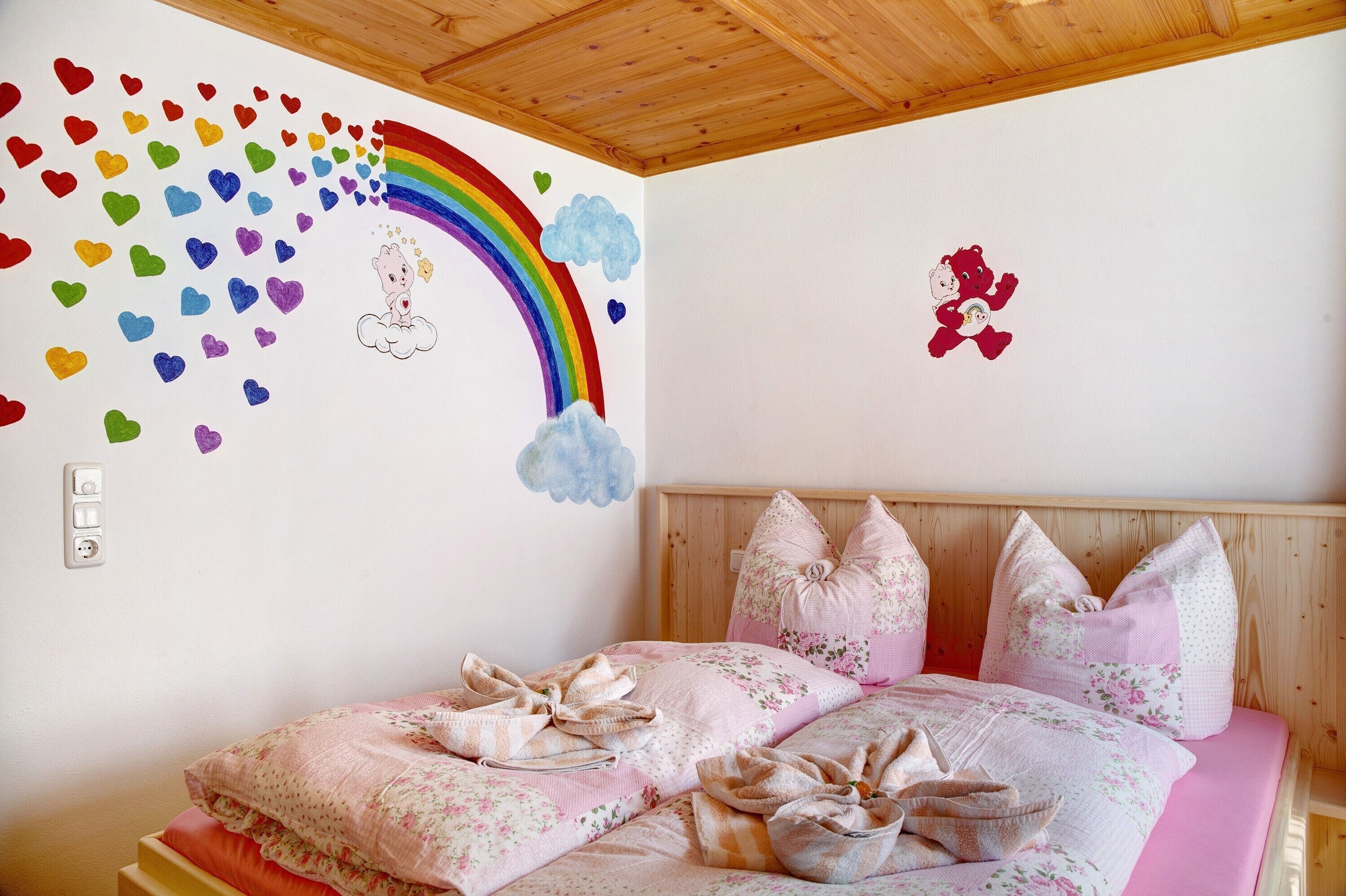 Een slaapkamer met een bed, kussens, een houten hoofdbord, een muur met een regenboog en harten, en een pluche beertje sticker.
