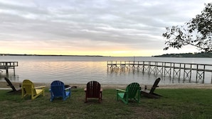 Blick auf ein Seeufer mit vier Adirondack-Stühlen, die zum Wasser zeigen, einer Holzsteg, der sich ins Wasser erstreckt, und einer Rasenfläche im Vordergrund.