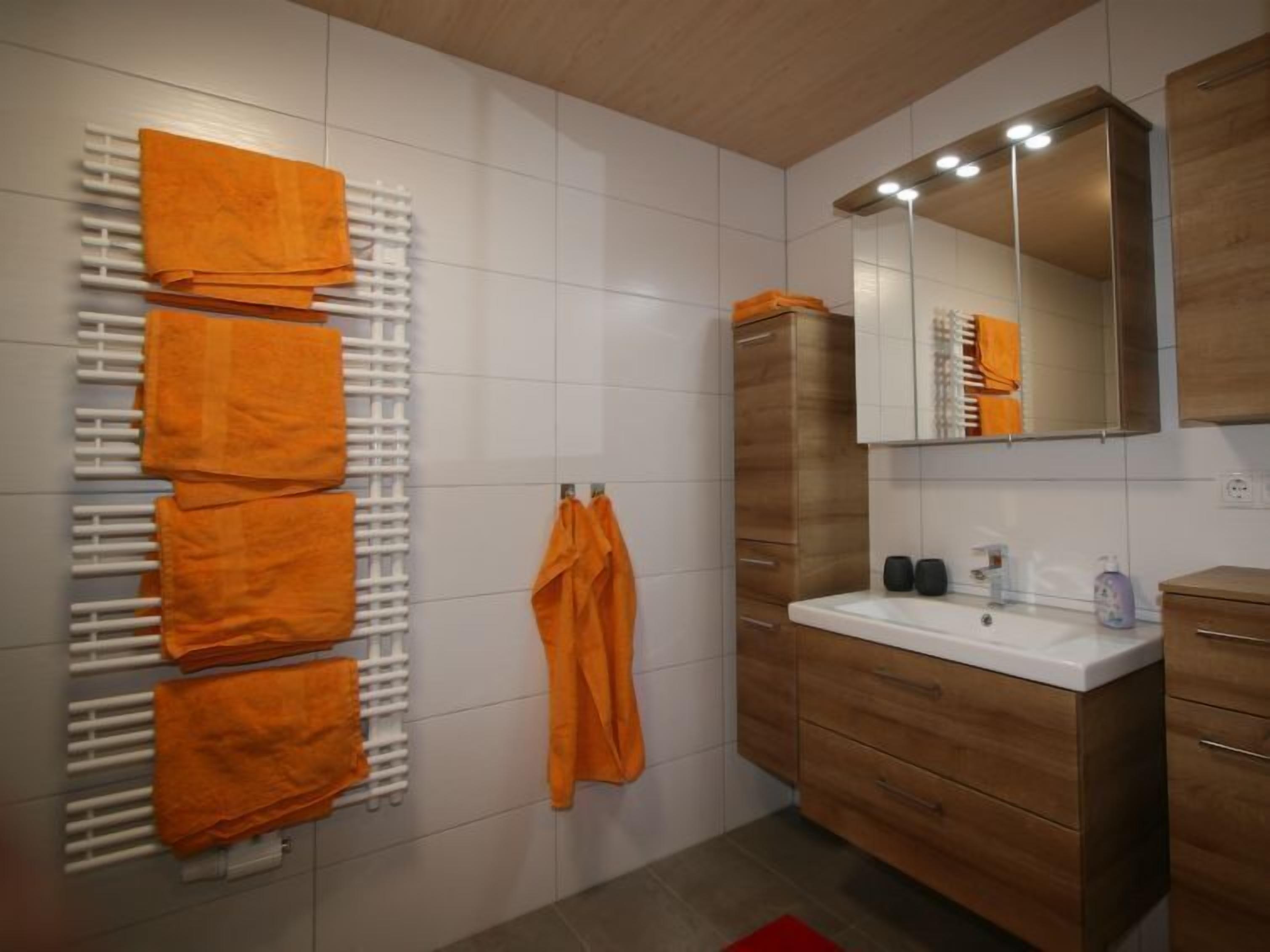 A modern bathroom with a white tiled wall, a wooden vanity with a mirror, and a towel rack with multiple orange towels.