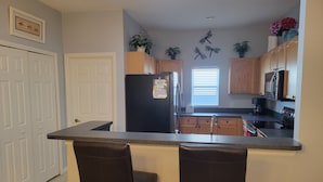 A kitchen with a black refrigerator, wooden cabinets, a black countertop, two bar stools, a window with blinds, and decorative butterfly wall art. - Magical Family Friendly Pool and Lake House (Kissimmee)